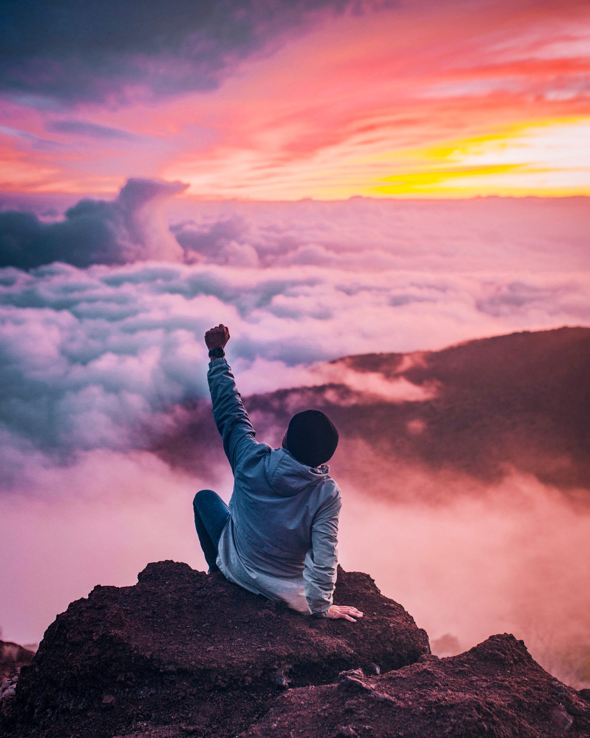 Man with Arm to sky in victory sitting at peak of mountain over hanging rock